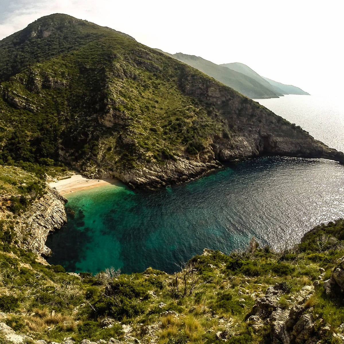 Aerial view of Dafina Bay & National Park Hidden Gems in Vlora, displaying turquoise waters surrounded by lush, rugged hills. A cozy sandy beach edges the water as a boat glides peacefully beneath a partly cloudy sky.