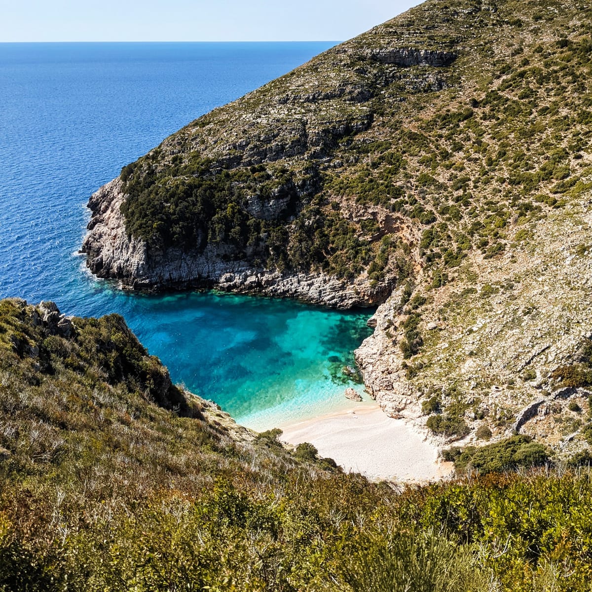 Bordered by rocky green hills, Dafina Bay & National Park Hidden Gems offers a secluded cove near Vlora with clear turquoise waters. The sandy beach, surrounded by steep cliffs, provides stunning views of the open sea—ideal for relaxing visits or boat excursions under a bright blue sky.