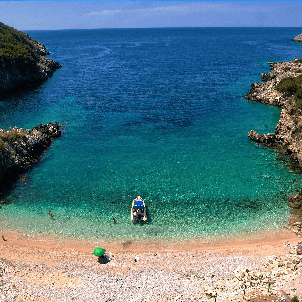 Discover Dafina Bay & National Park Hidden Gems: a tranquil beach with turquoise waters and rocky cliffs. A small boat awaits by the shore, where visitors swim and unwind. A green umbrella stands on the sandy coast beneath a clear blue sky.
