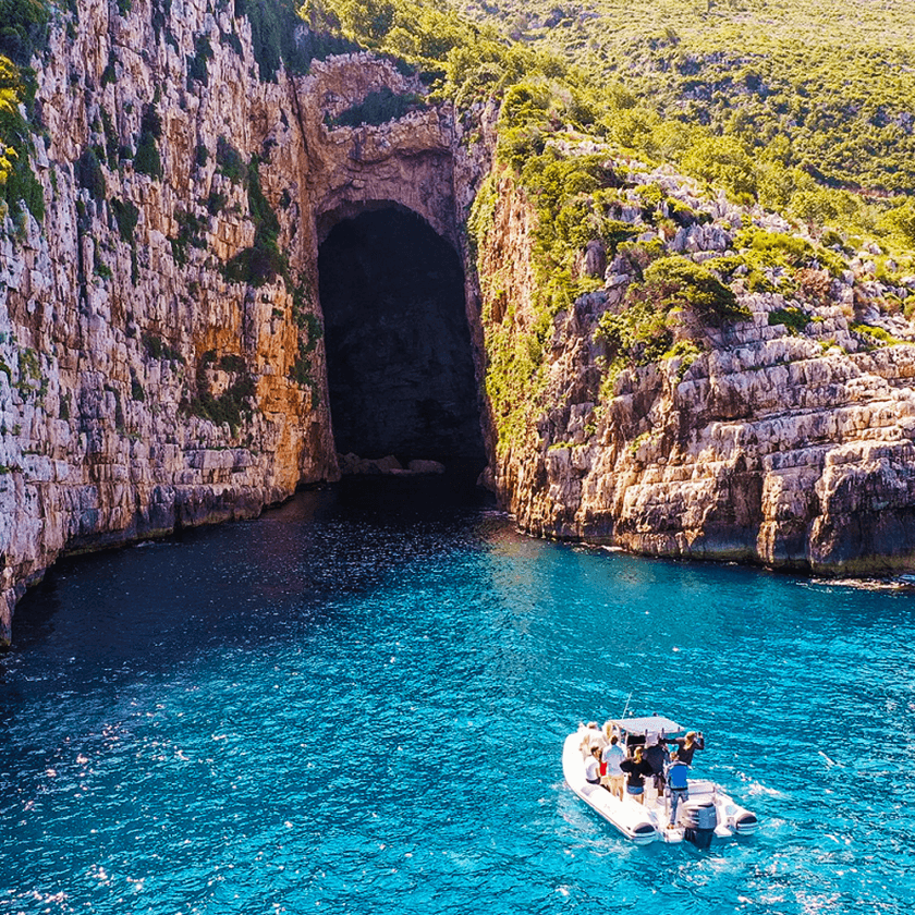 A small boat glides through the vibrant blue sea, offering trips to a large, dark cave entrance nestled in the rugged cliffs of Vlora. Sunlight dances on the water as lush greenery crowns the rocky heights above.