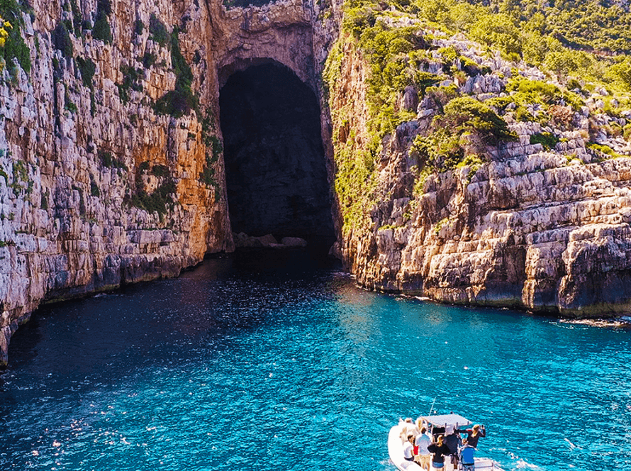 A small boat glides through the vibrant blue sea, offering trips to a large, dark cave entrance nestled in the rugged cliffs of Vlora. Sunlight dances on the water as lush greenery crowns the rocky heights above.