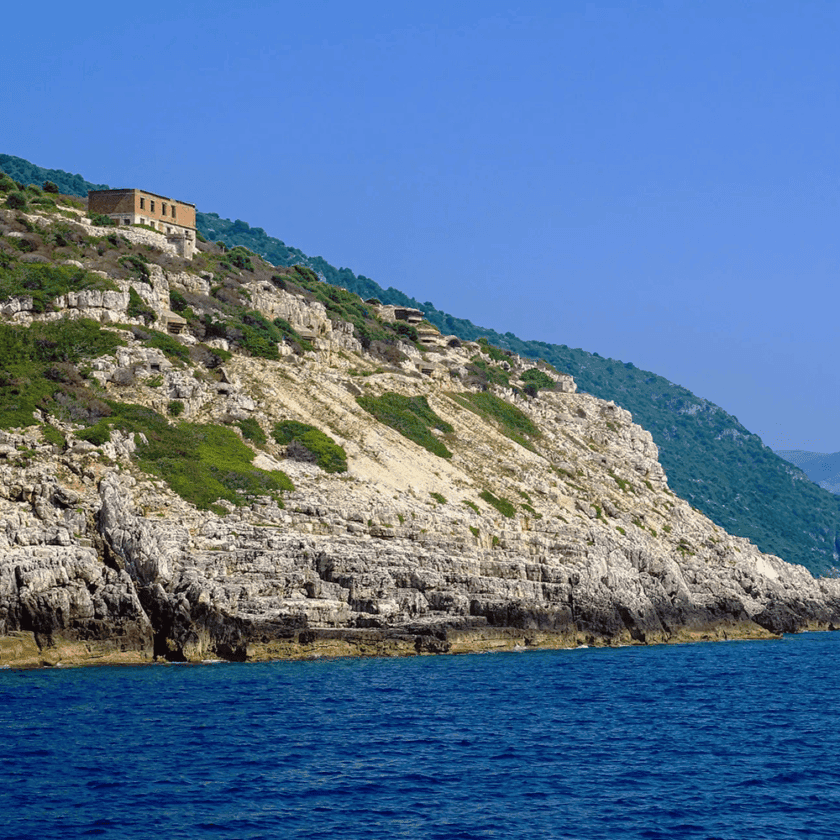 Rocky coastal landscape near Vlora with a blue sea in the foreground. A rustic, abandoned building sits atop the steep, green-covered cliffs under a clear blue sky. The scene, ideal for boat trips, is bathed in bright sunlight.