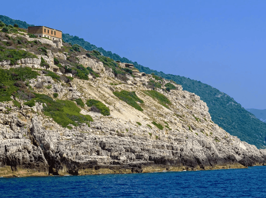Rocky coastal landscape near Vlora with a blue sea in the foreground. A rustic, abandoned building sits atop the steep, green-covered cliffs under a clear blue sky. The scene, ideal for boat trips, is bathed in bright sunlight.