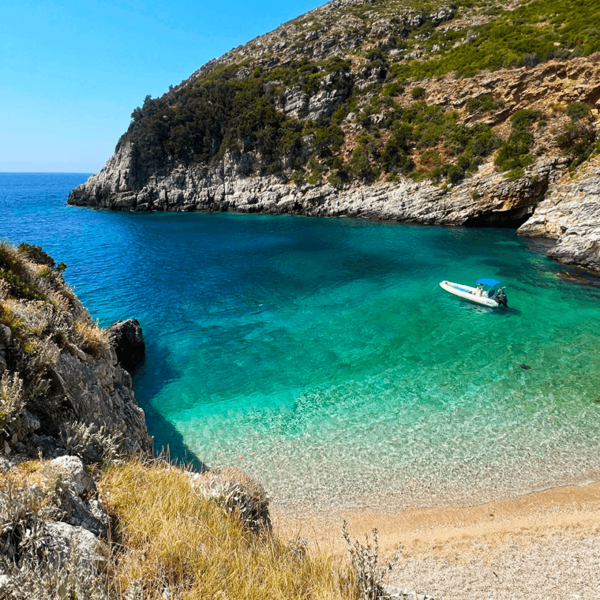 A small, white motorboat from Vlora floats on the clear turquoise waters of a secluded cove, surrounded by rugged cliffs and lush greenery. The shoreline features a narrow, sandy beach, perfect for day trips under the clear blue sky.