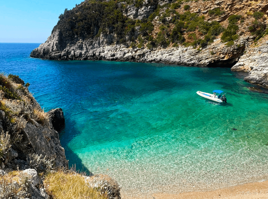 A small, white motorboat from Vlora floats on the clear turquoise waters of a secluded cove, surrounded by rugged cliffs and lush greenery. The shoreline features a narrow, sandy beach, perfect for day trips under the clear blue sky.