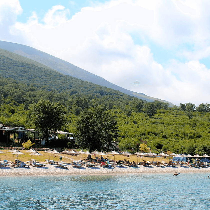 The beach at Vlora offers sun loungers and umbrellas along the shoreline, where people relax under the shade. In the background, green hills and trees frame a partly cloudy sky, perfect for setting sail on nearby boat trips.
