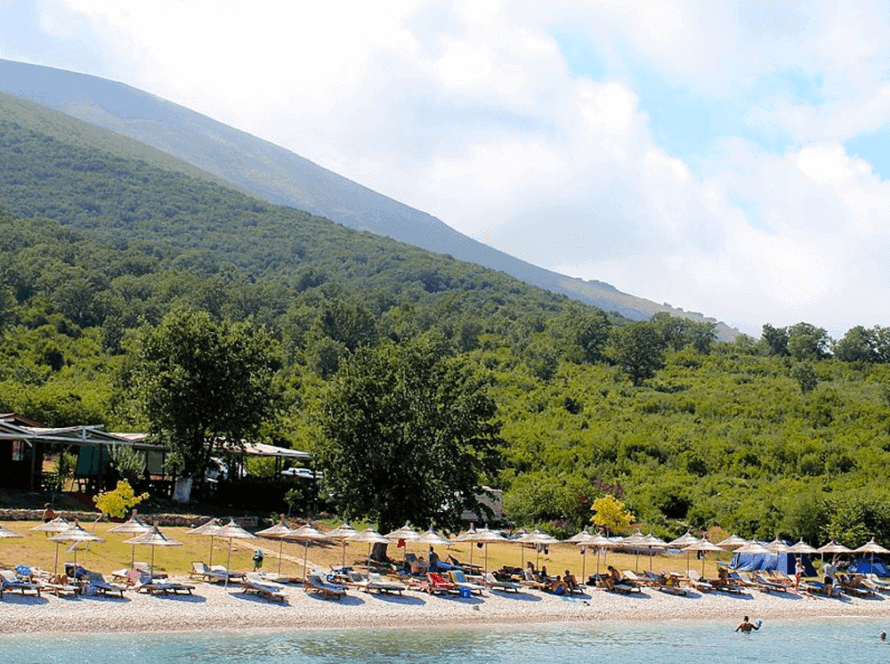 The beach at Vlora offers sun loungers and umbrellas along the shoreline, where people relax under the shade. In the background, green hills and trees frame a partly cloudy sky, perfect for setting sail on nearby boat trips.