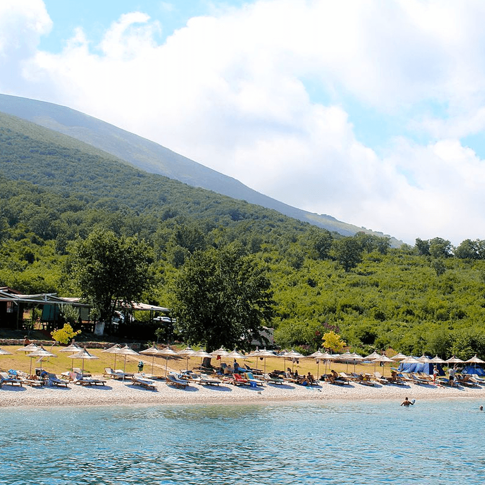 The beach at Vlora offers sun loungers and umbrellas along the shoreline, where people relax under the shade. In the background, green hills and trees frame a partly cloudy sky, perfect for setting sail on nearby boat trips.