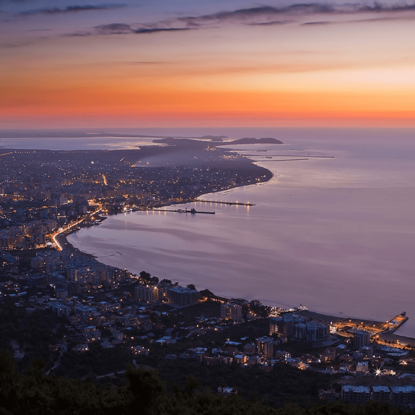 Coastal city at dusk with buildings and streets illuminated. The shoreline curves gracefully as boats gently sway, and distant hills are visible under a colorful sky transitioning from deep orange to blue. The sea reflects the evening light, creating a serene atmosphere perfect for memorable trips.