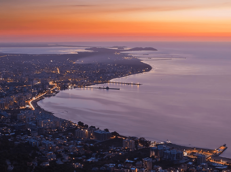 Coastal city at dusk with buildings and streets illuminated. The shoreline curves gracefully as boats gently sway, and distant hills are visible under a colorful sky transitioning from deep orange to blue. The sea reflects the evening light, creating a serene atmosphere perfect for memorable trips.