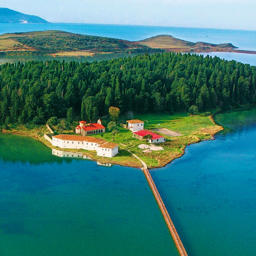Aerial view of a small green island near Vlora with a cluster of red-roofed buildings, surrounded by blue water. A long bridge and trips on boats connect the island to the mainland. Forested areas and distant hills are visible in the background.