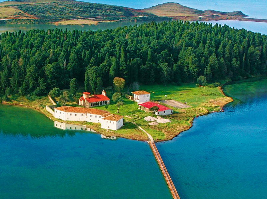 Aerial view of a small green island near Vlora with a cluster of red-roofed buildings, surrounded by blue water. A long bridge and trips on boats connect the island to the mainland. Forested areas and distant hills are visible in the background.