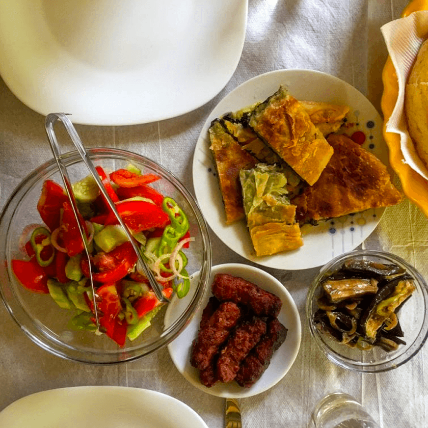 A table set with a fresh salad of tomatoes and peppers, a plate of savory pastries, a small dish of grilled sausage, pickled vegetables in a glass, and a covered tea set, all arranged on a white tablecloth.