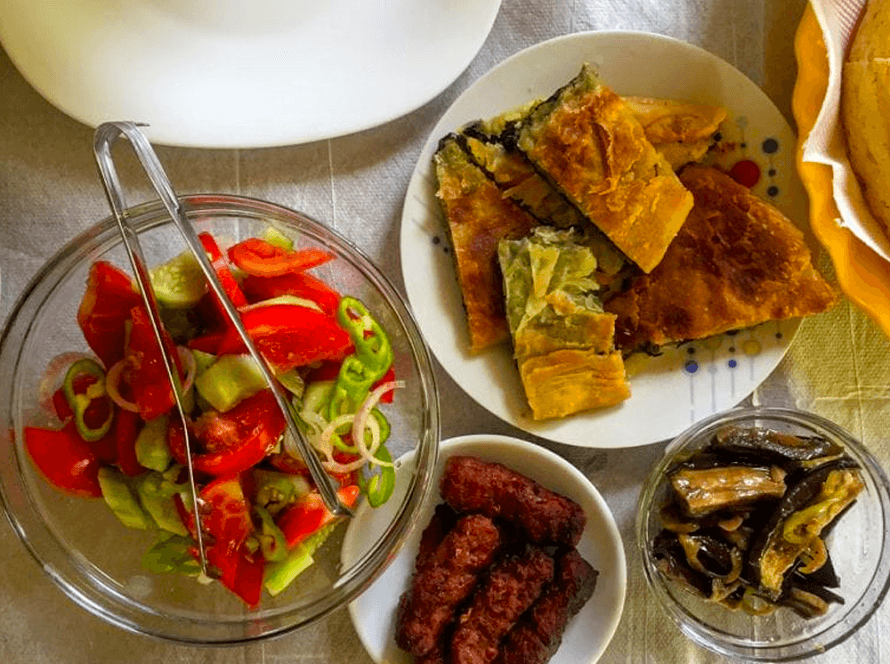 A table set with a fresh salad of tomatoes and peppers, a plate of savory pastries, a small dish of grilled sausage, pickled vegetables in a glass, and a covered tea set, all arranged on a white tablecloth.