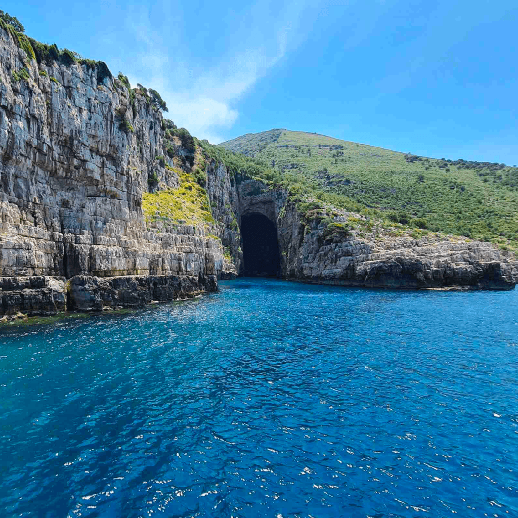 Crystal clear blue water surrounds the tall, rocky cliffs with lush greenery on Sazan Island. A large, dark entrance to Haxhi Ali Cave is prominently visible in the center, contrasting beautifully with the bright sky and sea, encapsulating a serene and picturesque coastal landscape at Karaburun.