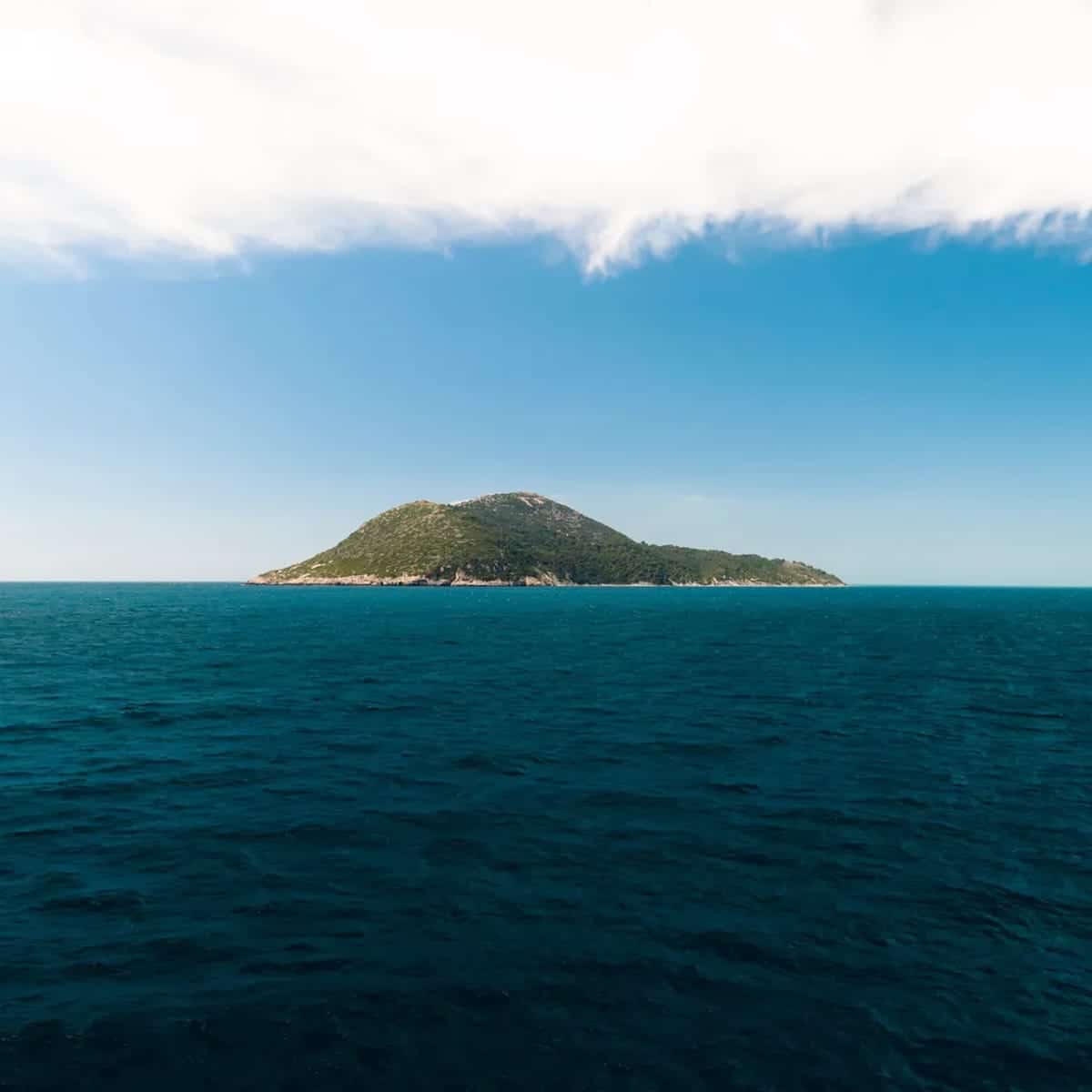 A small green island sits alone under a bright blue sky, surrounded by calm, deep blue ocean waters. A few wispy white clouds float above. Nearby, a boat sways gently, hinting at trips to the serene beauty of this hidden gem skirting Vlora.