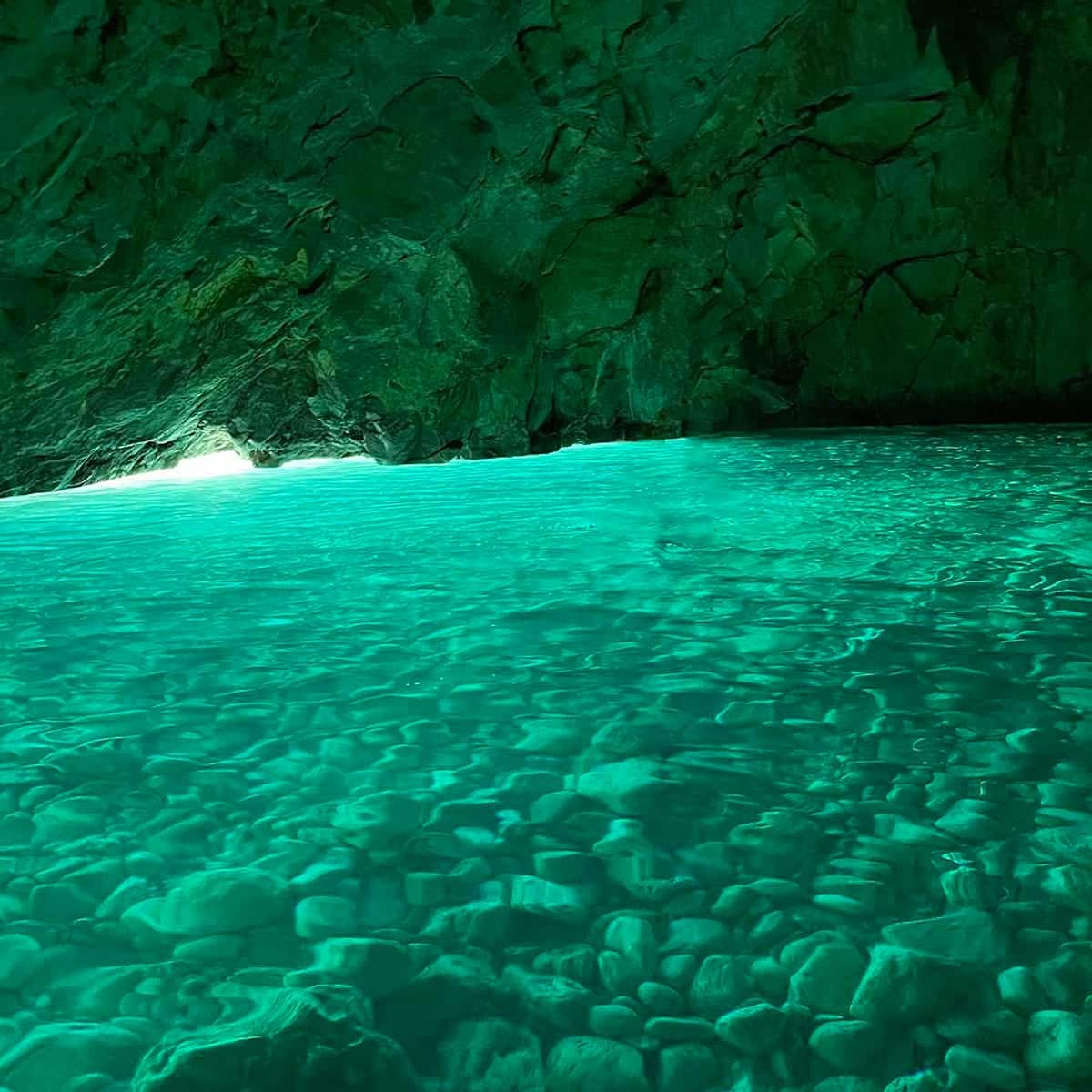 The Grama Bay Blue Cave and Marine Park features clear turquoise water, rocky walls, and sunlight filtering through openings, illuminating stones beneath the surface for a serene atmosphere.