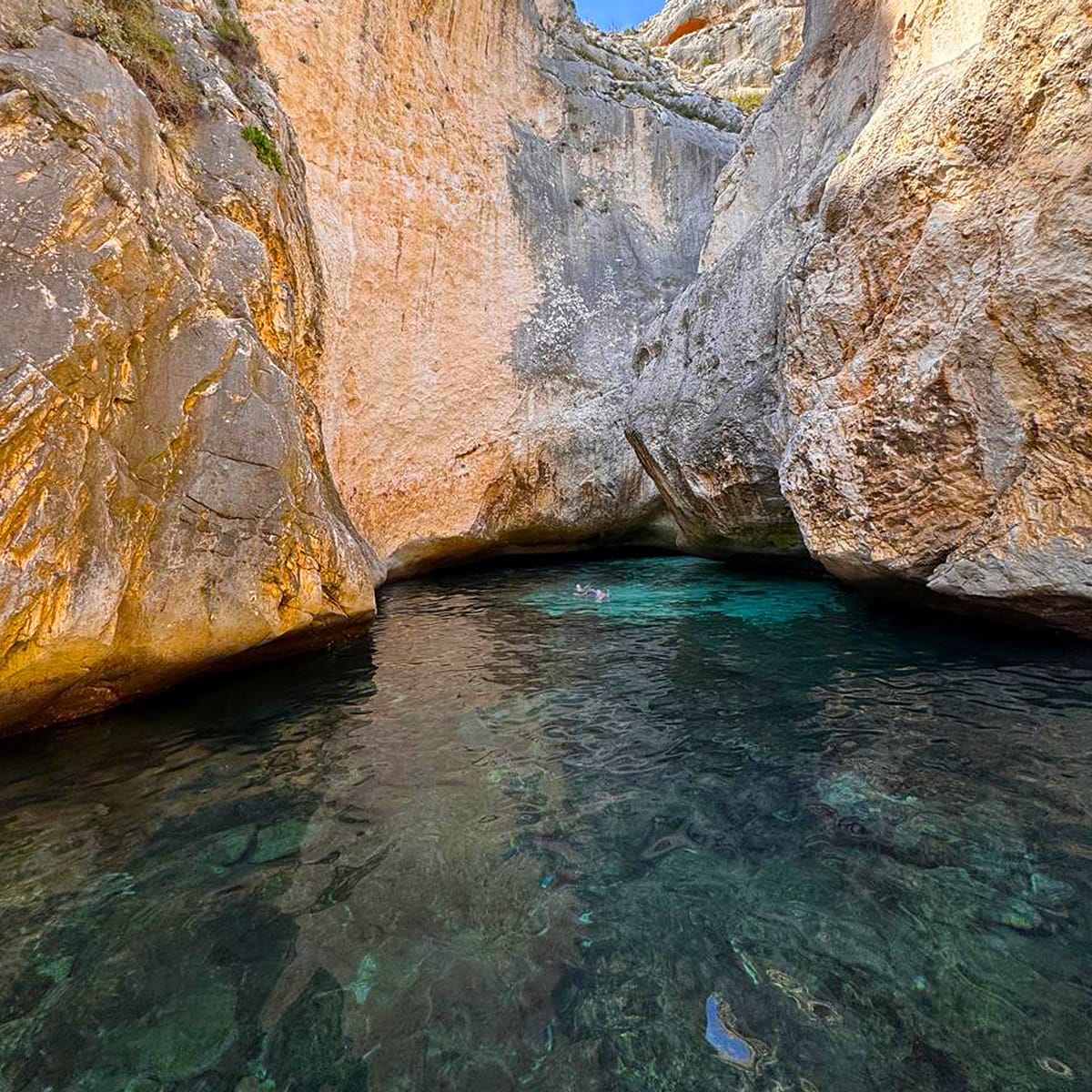 Grama Bays turquoise water flows through a rocky canyon, bordered by high cliffs. Sunlight casts a warm glow on the stone walls, and patches of blue sky peek above. Shadows add depth to the waters surface in this picturesque Blue Cave and Marine Park setting.